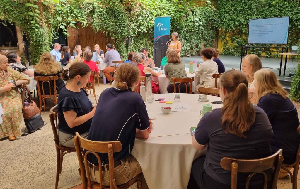A big room with lots of people sitting around round tables and listening to a facilitator at the front